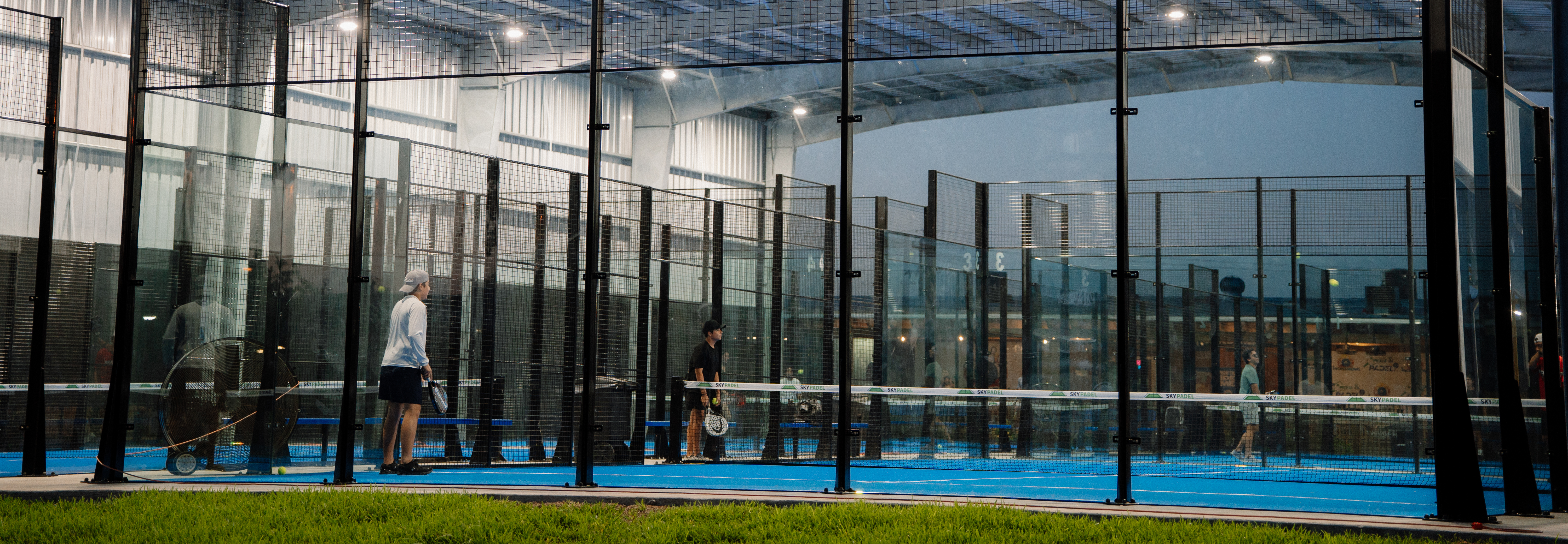 Padel
players rallying inside a glass-enclosed court on blue turf, dynamic doubles action
